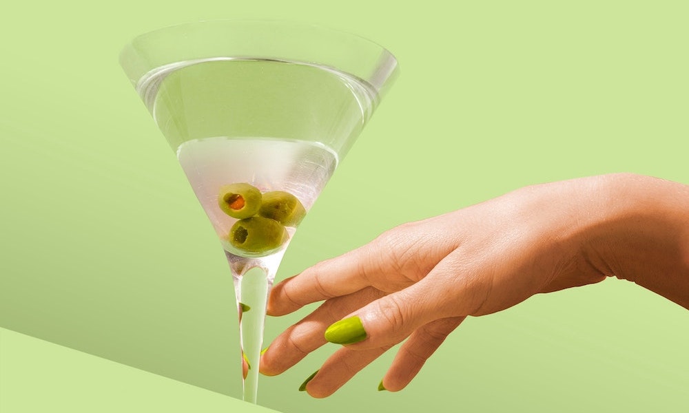Martini Glas mit Oliven und einer Hand die zum Glas greift auf olivgrünem Hintergrund
